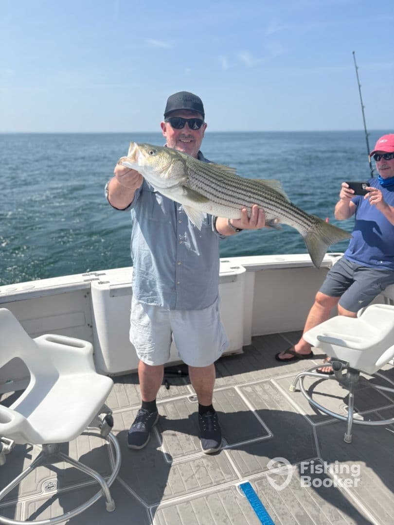 An angler proudly displaying a large Striped Bass aboard a fishing boat, enjoying a sunny day on the water.