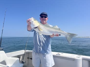 Angler proudly displaying a large Striped Bass while aboard a fishing boat in clear waters.