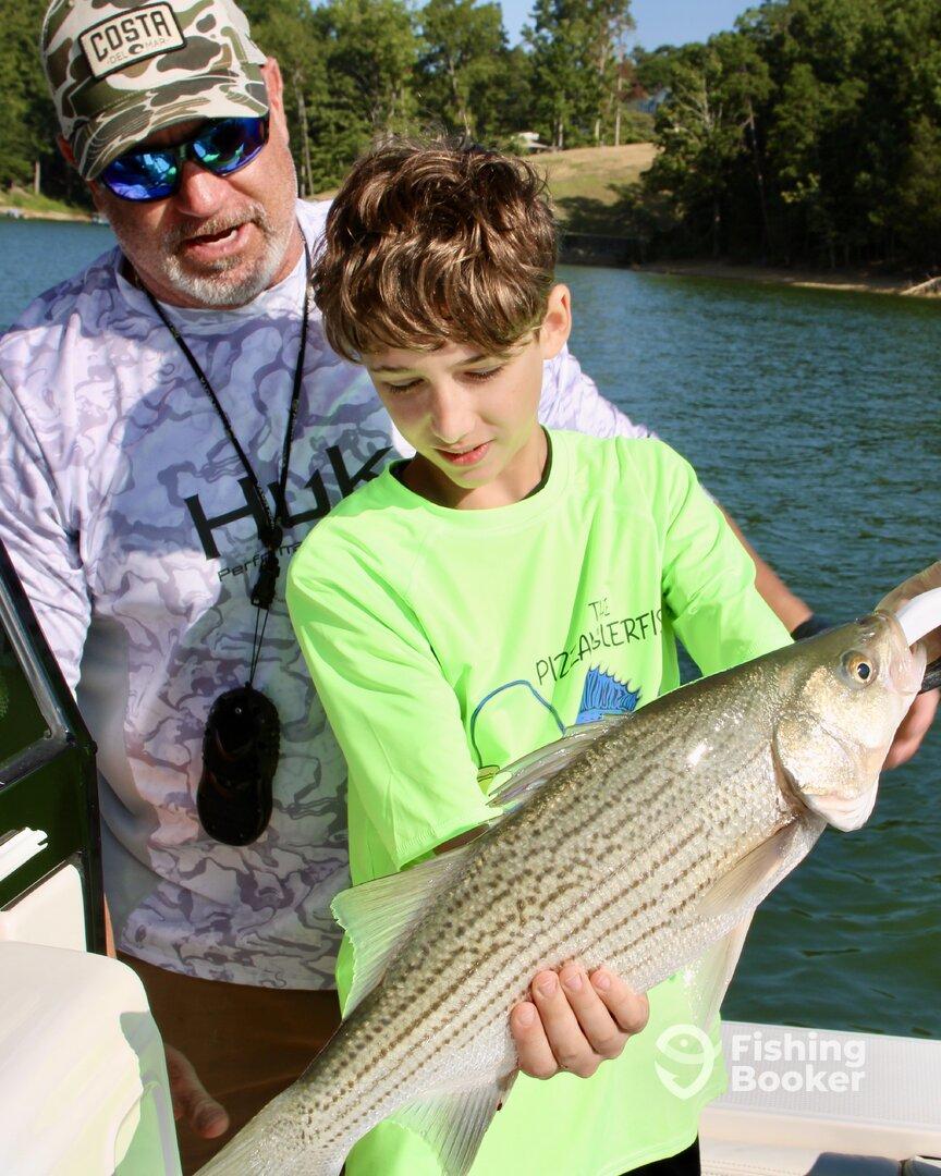 A young angler proudly displaying a striped bass while fishing on a lake, accompanied by an adult mentor.