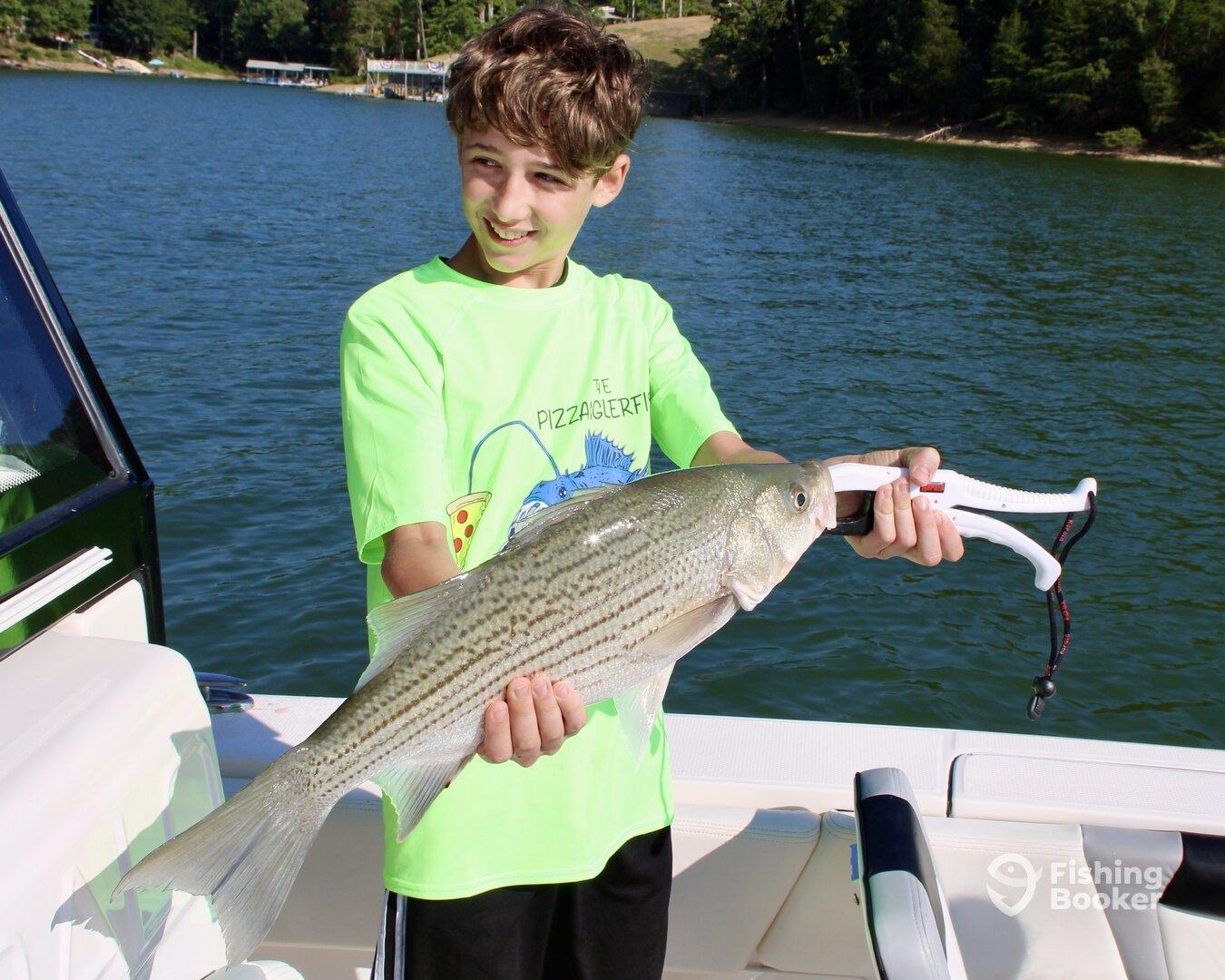 A young angler proudly displaying a striped bass while fishing on a lake, showcasing a vibrant green shirt against a scenic backdrop.