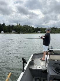 A young angler casting a line from a small aluminum boat on a serene lake, surrounded by lush greenery and cloudy skies.