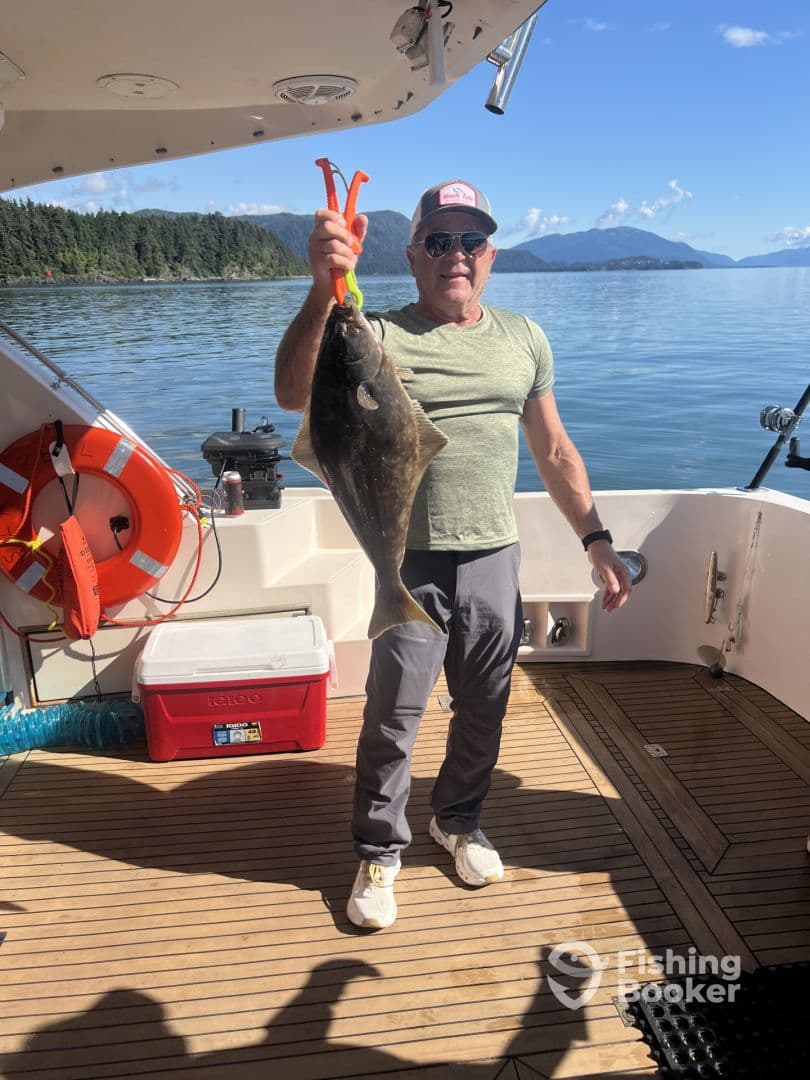 An angler proudly displaying a large Halibut while aboard a fishing boat in a scenic coastal environment.