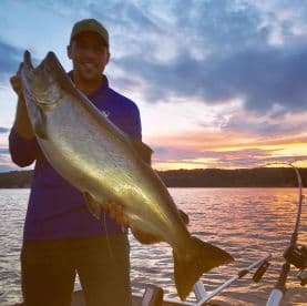Angler proudly displaying a large Salmon against a stunning sunset backdrop on a fishing trip.