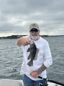 An angler proudly displaying a Black Sea Bass while fishing on a boat, showcasing a successful catch in a scenic coastal environment.