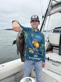 Angler proudly displaying a large Black Sea Bass while aboard a fishing boat, showcasing a successful catch during a cloudy day on the water.