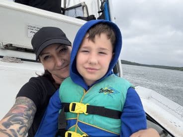 A woman and a young boy enjoying a moment on the boat, both wearing life jackets, with a scenic water background.