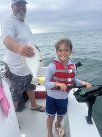 A young boy joyfully holds a Flounder while fishing on a boat, showcasing a memorable moment with an adult companion.
