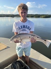 A young angler proudly displaying a Redfish caught while fishing from a boat in a scenic coastal environment.