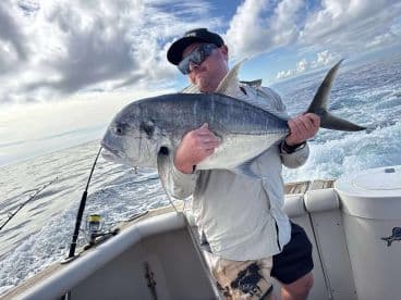 Angler holding a large Giant Trevally while fishing on a boat, showcasing a successful catch in open waters.