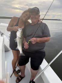 Two anglers proudly displaying a large Striped Bass while fishing on a boat, enjoying a memorable day on the water.