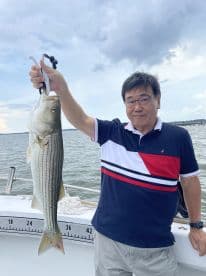 An angler proudly displaying a large Striped Bass while fishing on a boat, showcasing a successful day on the water.
