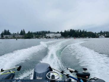 A scenic view from the back of a boat, showcasing the wake and the shoreline with buildings and trees, typical of a coastal fishing environment.