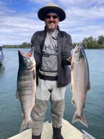 Angler proudly displaying two large Salmon caught during a fishing trip on a scenic river.