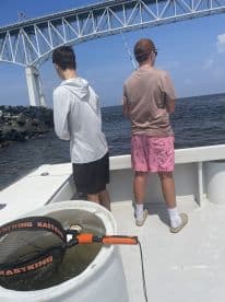 Two young anglers fishing off the side of a boat under a bridge, showcasing a lively fishing environment.
