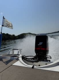 A view from the back of a boat showcasing the powerful engine and the flag of 'Old Questers' as it speeds across a serene lake.