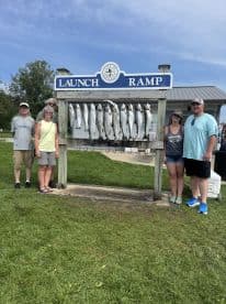 A group of anglers proudly posing in front of a catch board displaying multiple fish at a launch ramp, showcasing their successful fishing trip.
