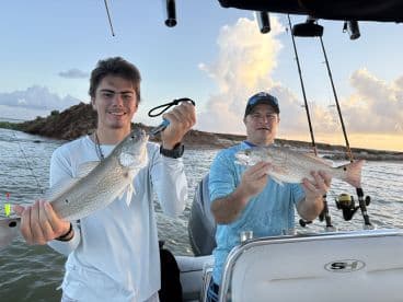 A young angler and an adult proudly displaying their catches of Redfish while fishing on a boat during sunset.