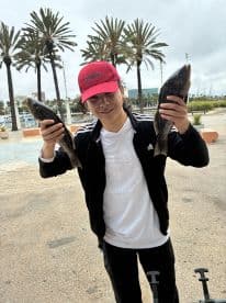 A young angler proudly displaying two caught fish in a scenic marina setting, surrounded by palm trees.