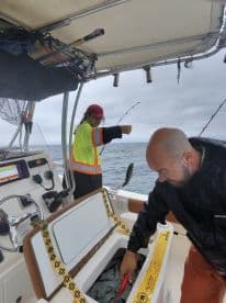A young angler is actively reeling in a fish while an adult prepares to store the catch in a cooler on a fishing boat, showcasing a dynamic fishing experience.