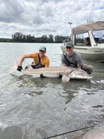 Two anglers proudly displaying a large Sturgeon while fishing from a boat in a river setting.