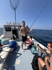 A young man is actively fishing on a boat, showcasing a catch while enjoying a sunny day at sea.