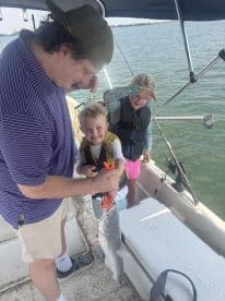 A young boy joyfully holds a caught fish with the help of an adult, while a girl looks on, all aboard a boat in a sunny fishing environment.