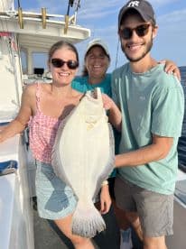 A group of three anglers proudly displaying a large Flounder aboard a fishing boat, enjoying a successful day on the water.
