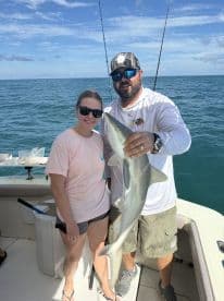 A young angler and an adult proudly displaying a caught shark aboard a fishing boat in clear blue waters.
