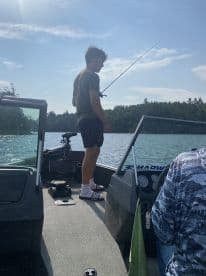 A young angler stands on a fishing boat, casting a line into a serene lake surrounded by lush greenery.