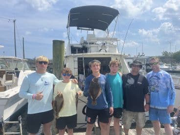 A group of anglers proudly displaying their catch of Flounder at the dock, with a fishing boat in the background.