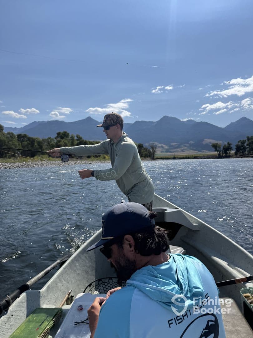 Two anglers casting lines from a small boat in a scenic river, surrounded by mountains, showcasing a peaceful fishing environment.