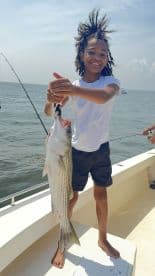 A young angler proudly holding a large Striped Bass while fishing on a boat, showcasing a successful day on the water.