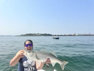 An angler proudly displaying a large Striped Bass while fishing on a sunny day, with a scenic backdrop of the coastline.