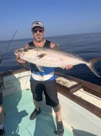 An angler proudly displaying a large Amberjack while fishing on a boat in calm waters.