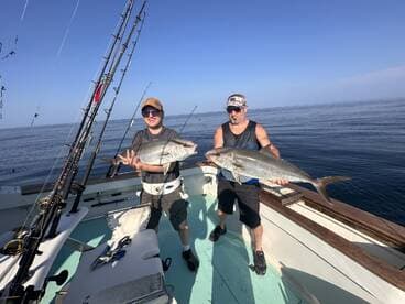 Two anglers proudly displaying their catches, a large Amberjack and a Snapper, aboard a fishing boat in calm waters.