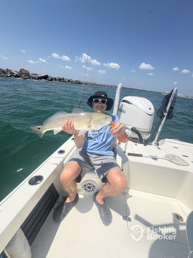Angler proudly displaying a large Redfish while seated on a fishing boat, with a scenic coastal backdrop.