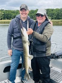 Two anglers proudly displaying a large Northern Pike while fishing on a serene lake.