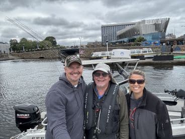 Three anglers posing on the deck of a fishing boat, showcasing the scenic waterfront and modern architecture in the background.