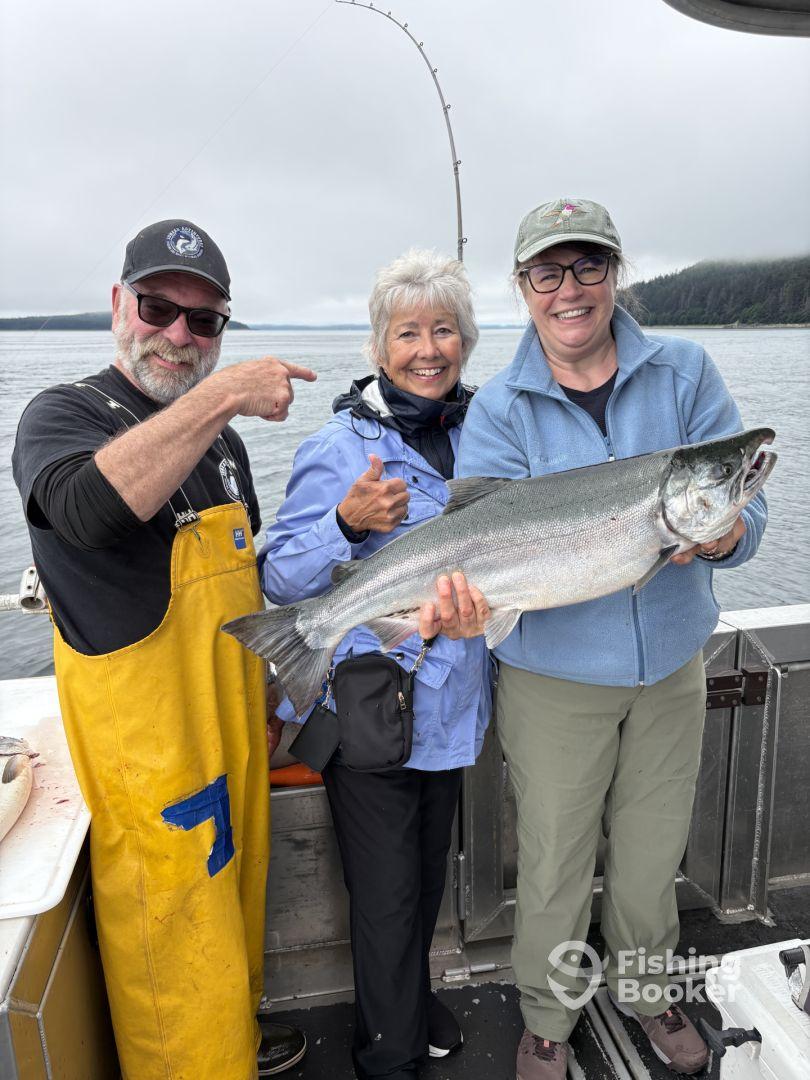 Three anglers celebrating a successful catch of a large Salmon while fishing on a boat in a scenic coastal environment.