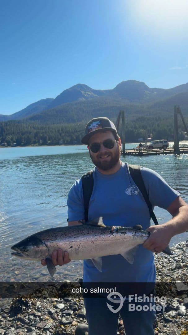 Angler proudly displaying a Coho Salmon against a scenic backdrop of mountains and water, showcasing a successful fishing trip.