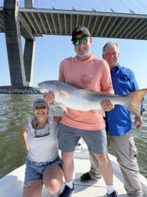 Three anglers proudly displaying a large Redfish on a boat under a bridge, enjoying a successful day of fishing.