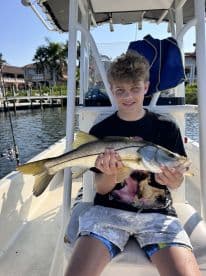 Young angler proudly holding a Snook while sitting on a boat in a scenic waterfront setting.