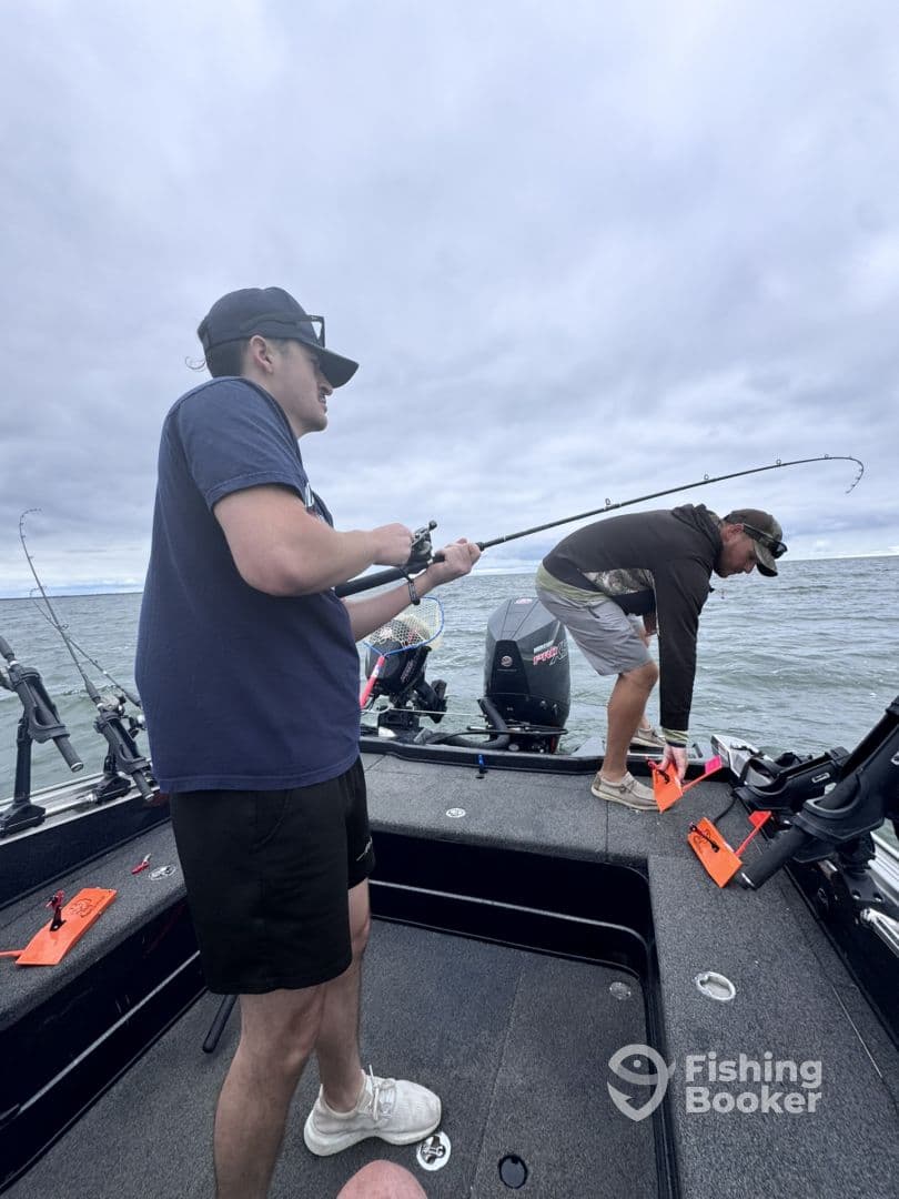 Two anglers actively fishing on a boat, with one reeling in a catch while the other prepares gear. The scene captures the excitement of a fishing trip on open water.