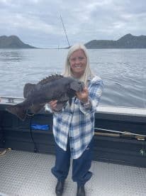 A proud angler holding a large Black Rockfish while fishing in a scenic coastal environment.