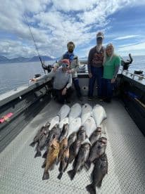 A group of four anglers proudly displaying their catch of various fish, including halibut and rockfish, on the deck of a fishing boat in a scenic coastal environment.