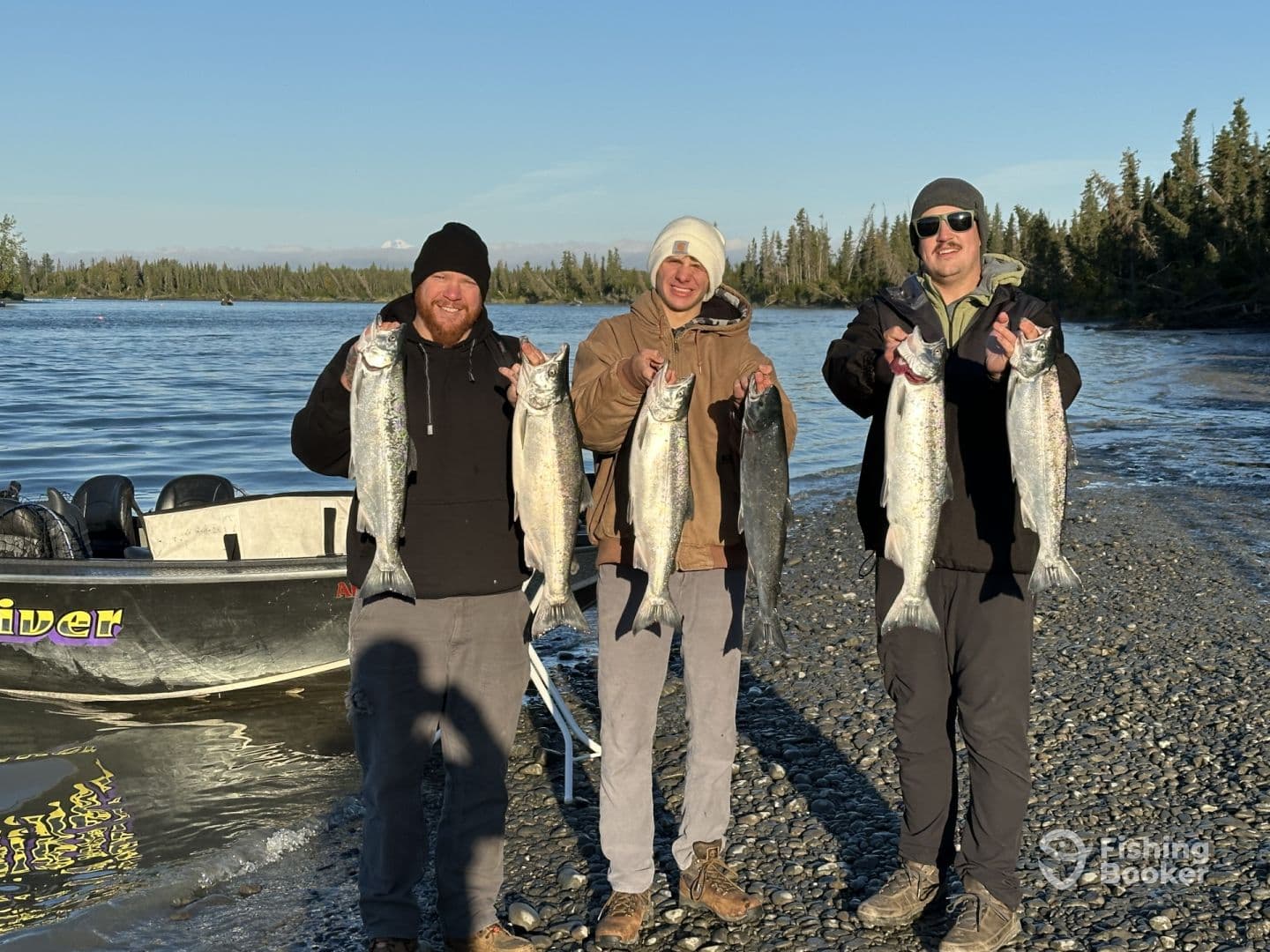 Three anglers proudly displaying their catch of salmon along a riverbank, showcasing a successful fishing trip.