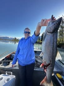 An angler proudly displaying a large Salmon while fishing on a serene lake, surrounded by lush greenery and mountains.