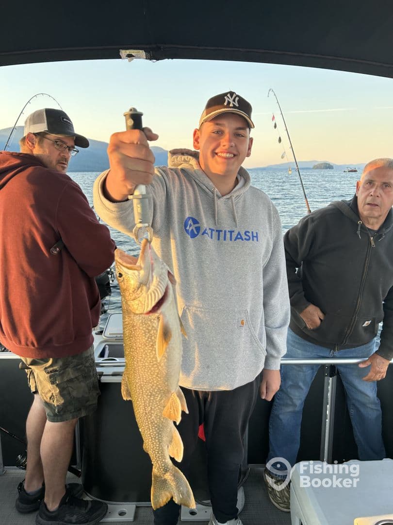 A young angler proudly displaying a large Lake Trout while fishing on a boat, with scenic views in the background.