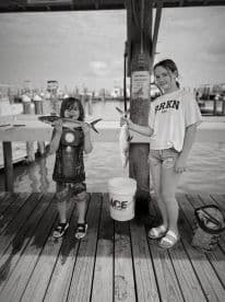 Two young anglers proudly displaying their catches, a small fish each, on the dock with a scenic marina backdrop.