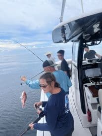 A group of anglers actively fishing on a boat, with one person reeling in a Red Snapper while others assist, showcasing a lively fishing experience.